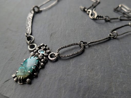 Flusskiesel inspirierte Silberkette Eine massive Silberkette mit einem Mittelteil in den ein Türkis und ein blauer Apatit eingefasst ist, liegt auf einem grauen Stein